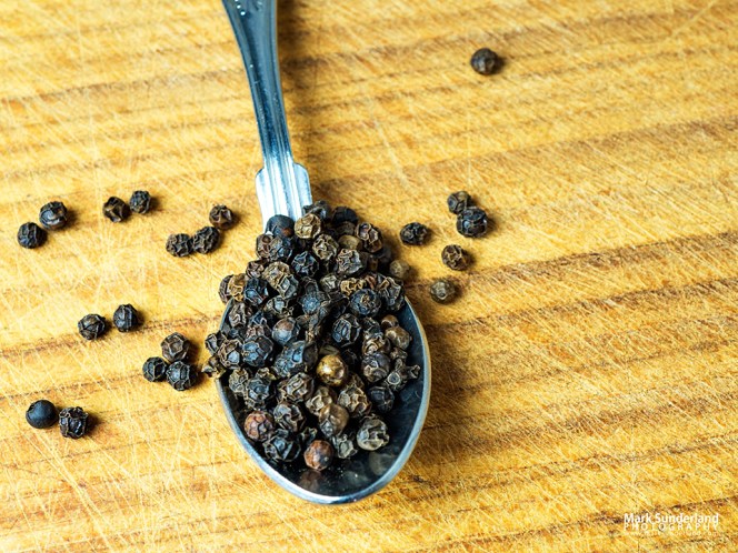 A teaspoonful of black peppercorns on a wooden chopping board with a few spilling over